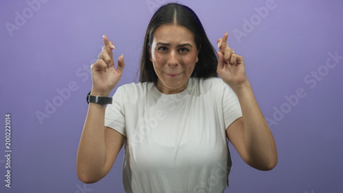 Foto Young brunette woman with crossed fingers and closed eyes in purple studio, hands near face and wearing white tee; hopeful anticipation