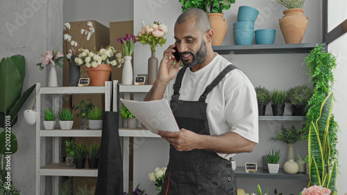 Photos Man florist holds smartphone to ear and reads order papers inside a shop building; focused order management