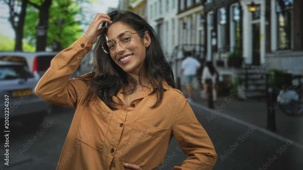 Obraz premium Woman touching hair with hand, smiling while wearing glasses and orange shirt, standing on street with parked cars and row houses; carefree joy.