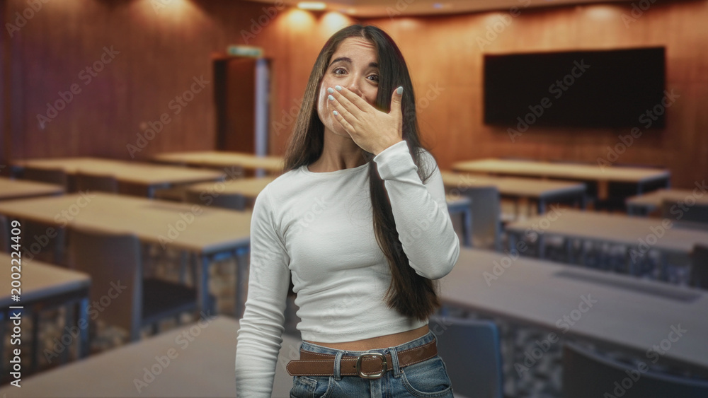 Fototapeta premium Young woman covers mouth with hand in a lecture building, standing among empty desks and a wall screen; surprise reflection.