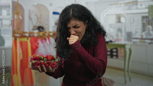 Foto Young woman holding plastic punnet of strawberries and coughing into her fist in building; allergy concern discomfort