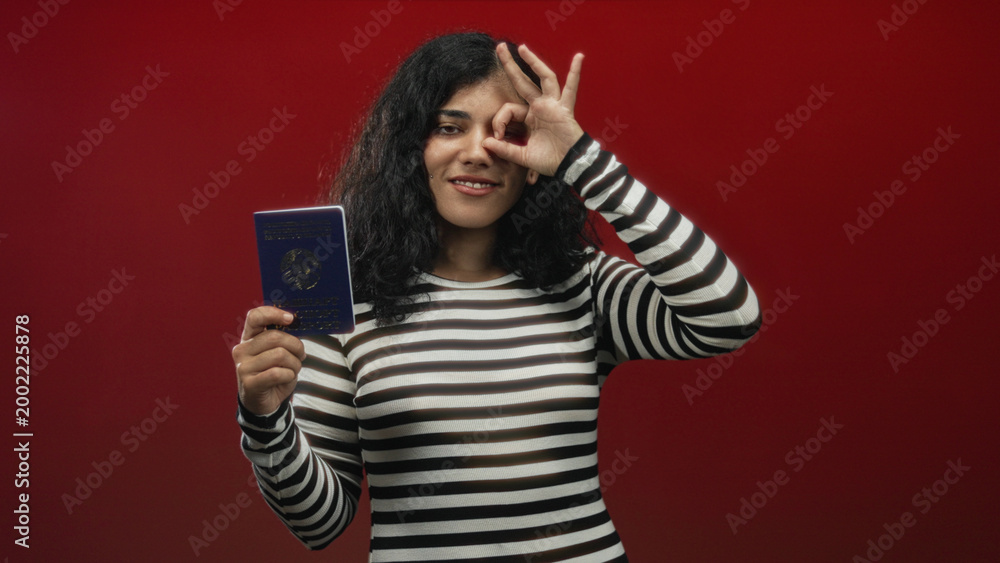 Naklejka premium Woman holding passport and making ok sign with hand in red studio while wearing striped top and smiling at camera; travel confidence.