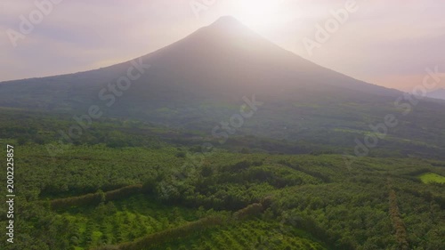 Aerial view of Volcano Arenal in Costa Rica during sunrise with sunlight illuminating the misty peak. Dense tropical rainforest covers the volcanic slopes and surrounding valley in lush green foliage.