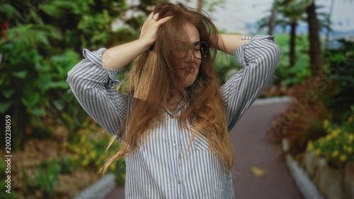 Fotografia Woman with glasses covers ears and holds head amid lush green forest pathway under sunlight; stress