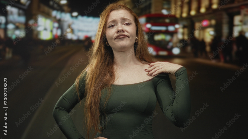 Fototapeta premium Redhead woman cups hand to ear on a busy city street at night near a red double decker bus and blurred crowd; frustration seeking help.