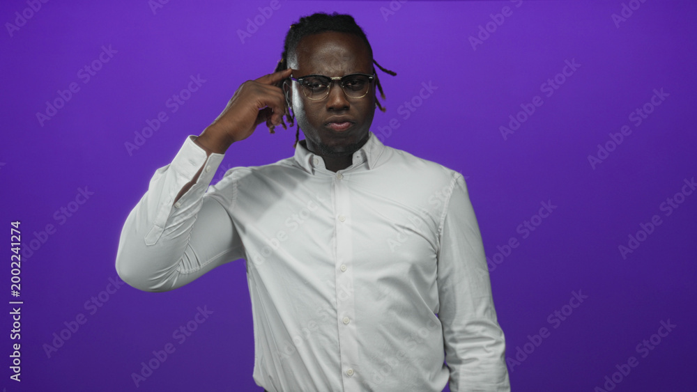 Naklejka premium Young african american man pointing finger to temple while wearing glasses and white shirt in studio purple backdrop; thoughtful.