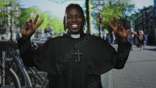 Φωτογραφία Man priest in cassock holds up peace sign with both hands on a busy street; joy faith community hope