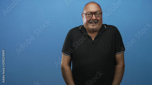 Photos Man, senior hispanic, standing and smiling with glasses and black polo, arms relaxed at sides against blue studio backdrop; cheerful confidence