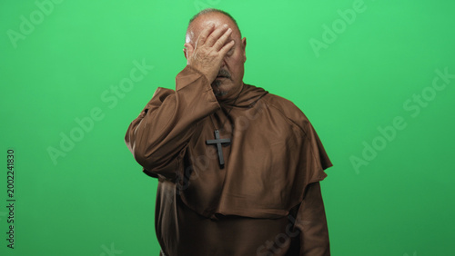 Quadro em tela Man in brown robe with hand covering face performing facepalm, wooden cross visible against green studio screen; remorse reflection