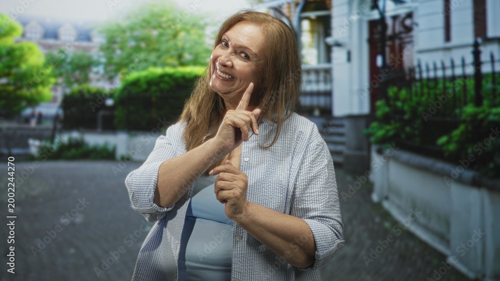 Fototapeta premium Woman smiling with hands pressed together and open palms in alternating gestures wearing light blue top and checked shirt on a residential street; gratitude joy.