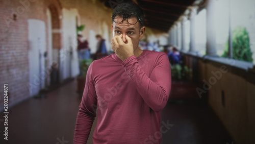Foto Young hispanic man pinching nose with glasses on head in a building corridor, covering mouth from a pungent smell; discomfort odor