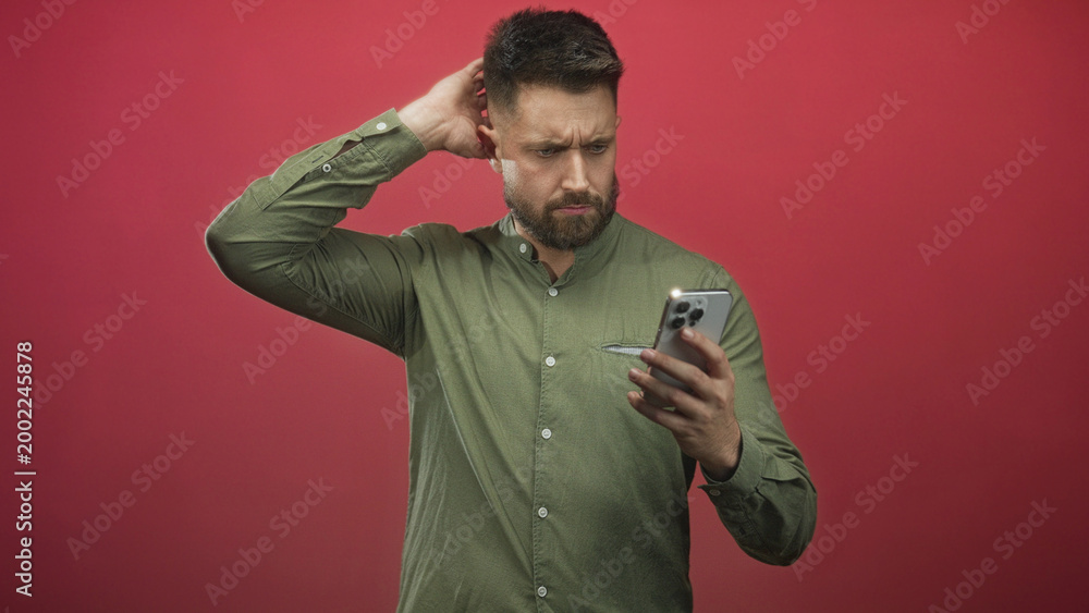 Fototapeta premium Man holding smartphone and touching ear while frowning and scratching his head, checking the screen in a red studio backdrop; confusion.