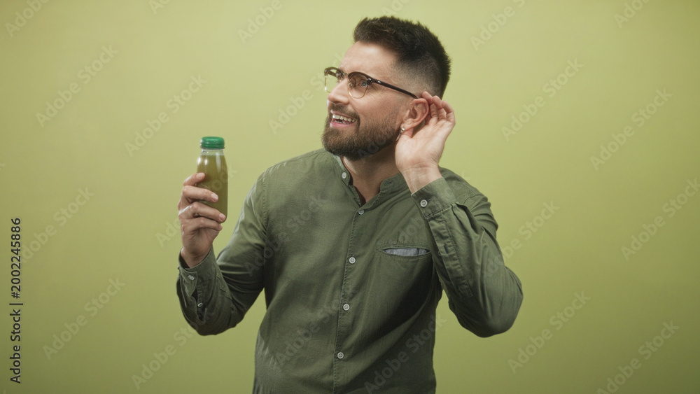 Naklejka premium Man with beard and glasses holding a bottled green juice with a green cap, cupping his ear as if listening while smiling in studio; curiosity wellness.
