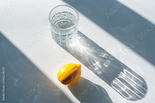 Tablou pe pânză Minimal still life with glass of water and fresh lemon casting sharp shadows on white table in sunlight