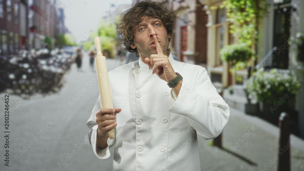Fototapeta premium Man chef in white uniform holding rolling pin and finger to lips for silence on street; quiet secrecy.
