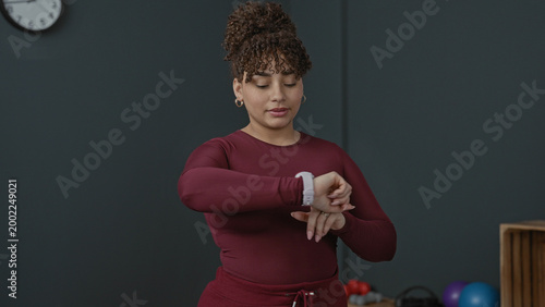 Fotografía Woman checking smartwatch on wrist and adjusting stopwatch strap in studio with exercise balls and clock visible; steady concentration