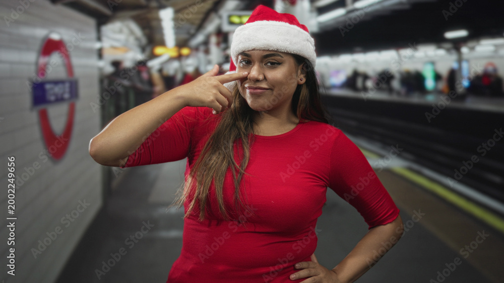 Obraz premium Woman wearing santa hat points finger to nose on a subway platform inside a building; holiday playful cheer.