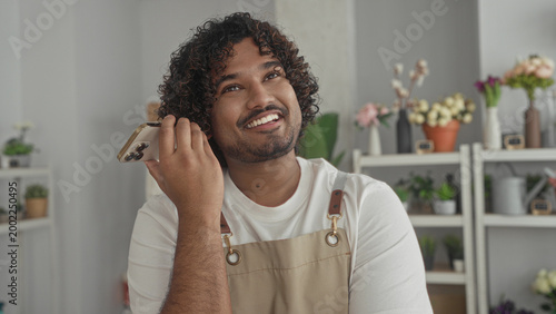 Man florist holding smartphone to ear and smiling in a studio shop wearing apron; small business joy.