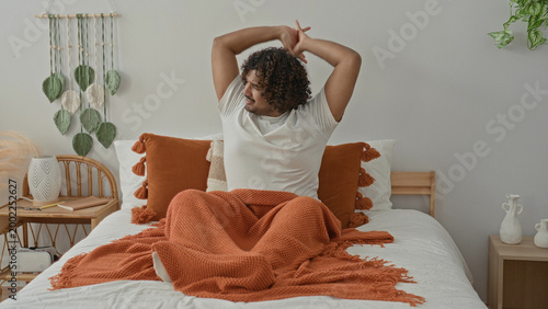 Foto Young indian man on bed stretches arms up, yawning with orange blanket, tassel pillow and wicker bedside table in studio; morning serenity