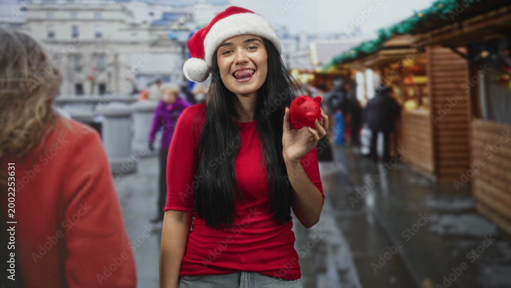 Naklejka premium Young woman holds red piggy bank, tongue out, wearing santa hat at street christmas market; playful holiday savings.