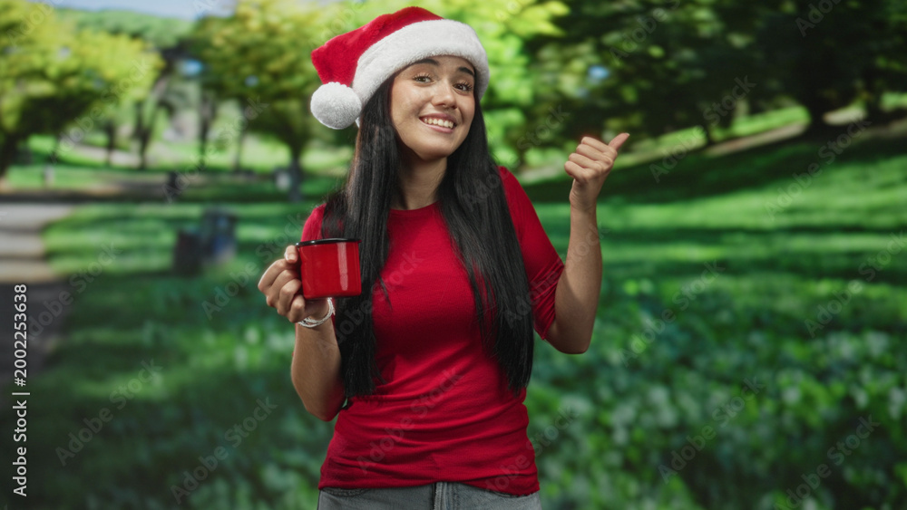 Naklejka premium Young woman wearing santa hat holding red mug and giving thumbs up in forest; holiday cheer cheerful.