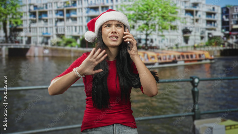Obraz premium Young woman in red santa hat holds phone to ear and grimaces, gesturing with hand beside a riverside building; frustration holiday stress.