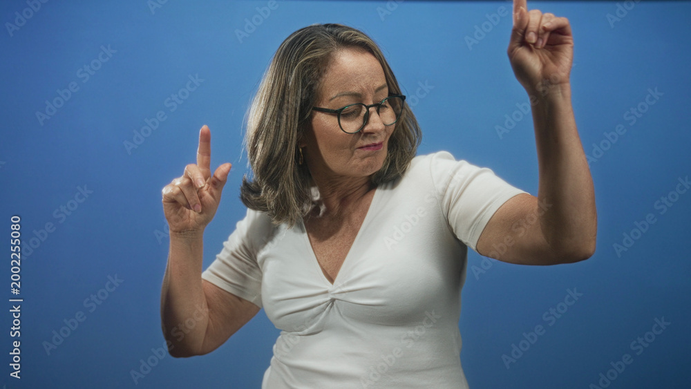 Fototapeta premium Hispanic senior woman wearing glasses and white shirt with raised hands and pointing finger dancing against blue studio backdrop; joyful celebration.