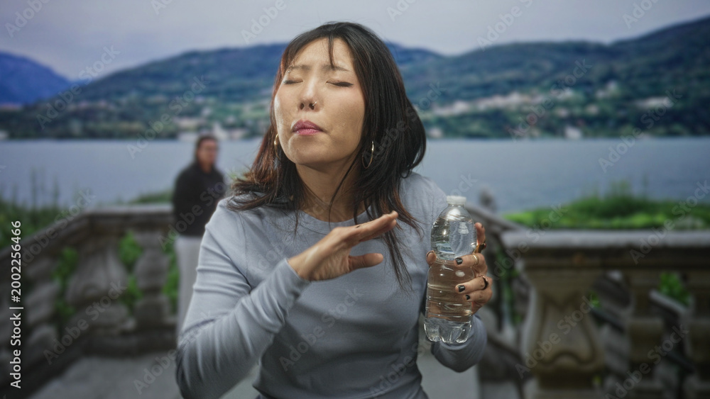 Naklejka premium Young woman holding water bottle, hand at throat, coughing on a building terrace by a lake; discomfort recovery.