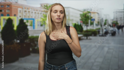 Woman with hand on chest showing breathlessness on a city street wearing black tank top and jeans; breath health relief.