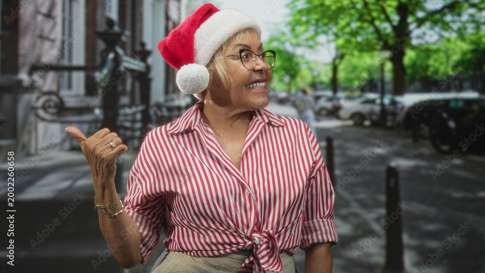 Fototapeta premium Woman wearing santa hat and glasses points thumb over shoulder on a leafy city street while smiling in a striped tied shirt and beige trousers; holiday joy.