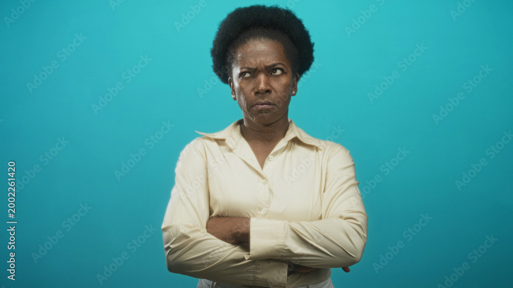 Fototapeta premium Woman with crossed arms and a frown standing centered in a studio portrait against a turquoise backdrop; disapproval resolve authority.
