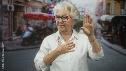 Canvas Print Senior woman with hand on chest and raised palm in busy market street, wearing white shirt and glasses, red nails visible; reflection dignity