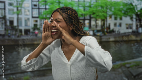 Фотография Woman face cups hands to mouth shouting on a street canal in amsterdam wearing a white blouse, braids visible; playful joy