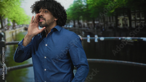 Foto Man hand to mouth calling on street in amsterdam by canal bridge, curly hair and blue shirt visible; urgent appeal