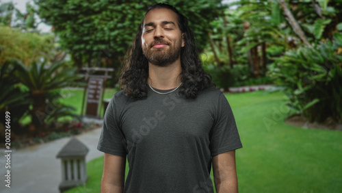 Foto Young man smiling with eyes closed and relaxed posture in a forest park near green lawn wearing gray tee; serenity mindfulness