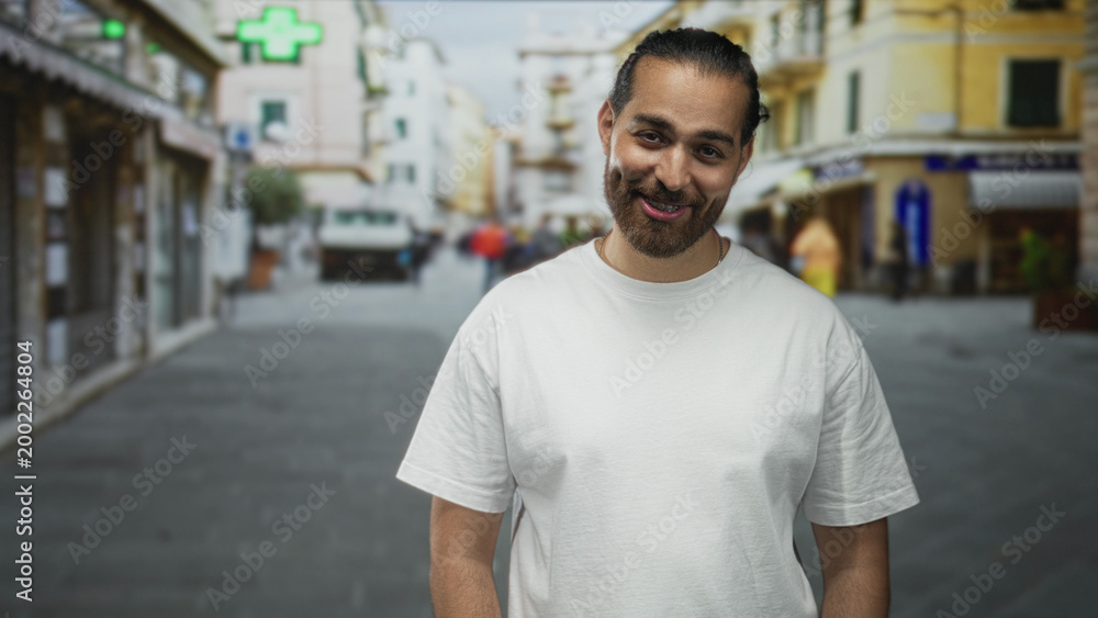 Fototapeta premium Man smiling in a white t shirt, standing on a busy city street with blurred shops and pedestrians; casual warmth.