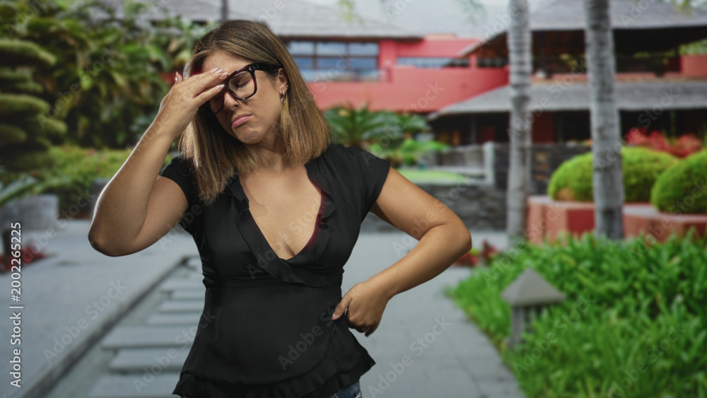 Fototapeta premium Woman touching temples and adjusting black glasses in hotel building courtyard, hands to head and squinting while grimacing; stress headache.