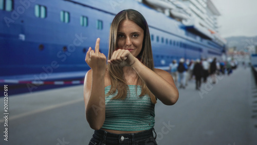 Schilderij op canvas Young woman shows middle finger and clenches fist at port beside a blue cruise ship, bare arms visible; defiance attitude