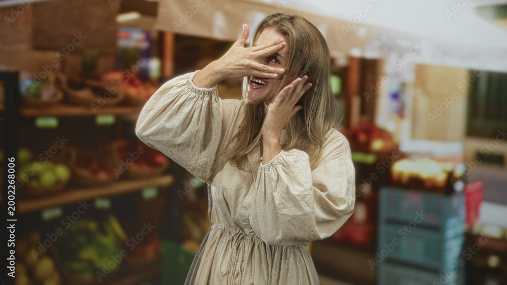 Fototapeta premium Blonde woman covers her face with a hand while smiling at a market stall in a building; playful surprise.