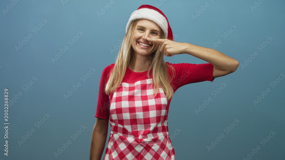 Naklejka premium Woman wearing santa hat and red checked apron points finger to nose while smiling in a studio with plain blue backdrop; playful holiday cheer.