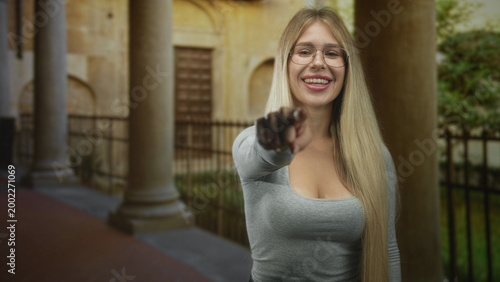 Free Download Young woman with glasses and long blonde hair showing cleavage points finger toward camera in a historic building colonnade; invitation confidence. - GFXLoad
