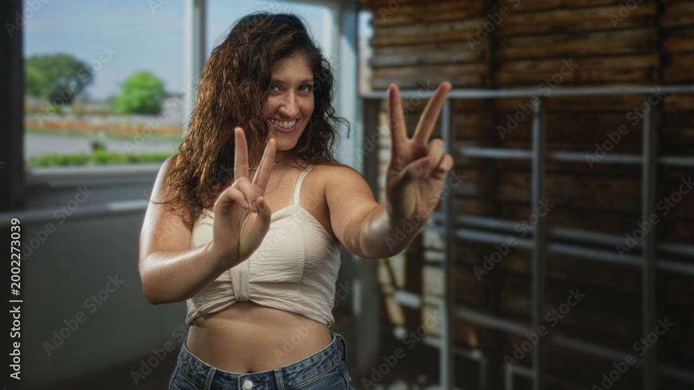 Fototapeta premium Woman showing double peace signs and bare midriff in studio space with window light and wooden rack; joy celebration.