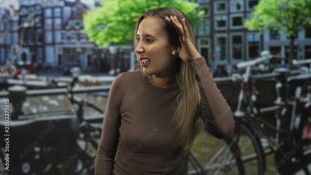 Fototapeta premium Woman young blonde with hand to hair beside bicycles along a canal on a street in amsterdam wearing brown top, gazing aside; quiet contemplation.