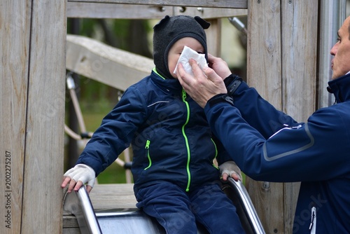 Parent wiping toddler’s nose with tissue on playground slide, caring outdoor moment showing protection, hygiene and family support during cold weather in everyday life scene.