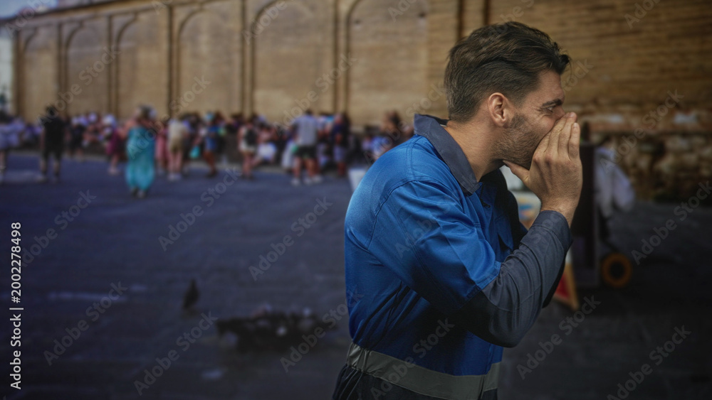 Obraz premium Man bearded worker in blue overalls cupping hands to mouth shouting toward a crowd on street by stone wall; crowd attention urgency.