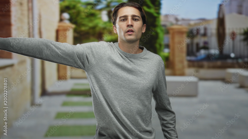 Fototapeta premium Man in gray sweater points finger and extends arm sideways on a sunlit stone street near benches and archway; curiosity decision.
