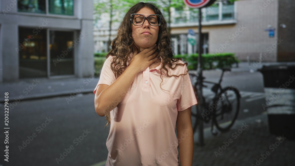Obraz premium Hispanic teenage woman holding hand to neck and wearing glasses on a street near a parked bicycle and building entrance; fatigue discomfort.