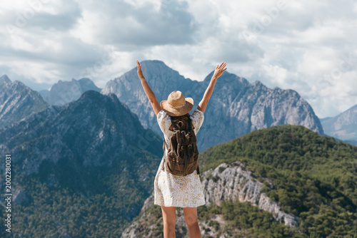 A rear view of a happy female tourist in a straw hat standing on a cliff edge with hands raised in the air. She is celebrating success and freedom while looking at the majestic mountain landscape.