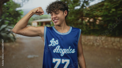 Canvas Print Teen boy flexes biceps and shows smile while wearing blue los angeles basketball jersey in street with stone wall and trees; youthful confidence