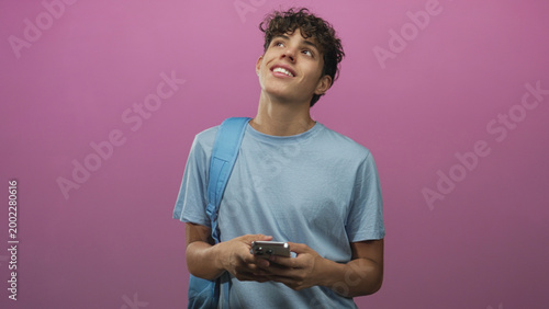 Konstfotografi Teen boy wearing light blue tee and backpack holding smartphone with bare hands, looking up and smiling in studio; daydreaming thoughtful